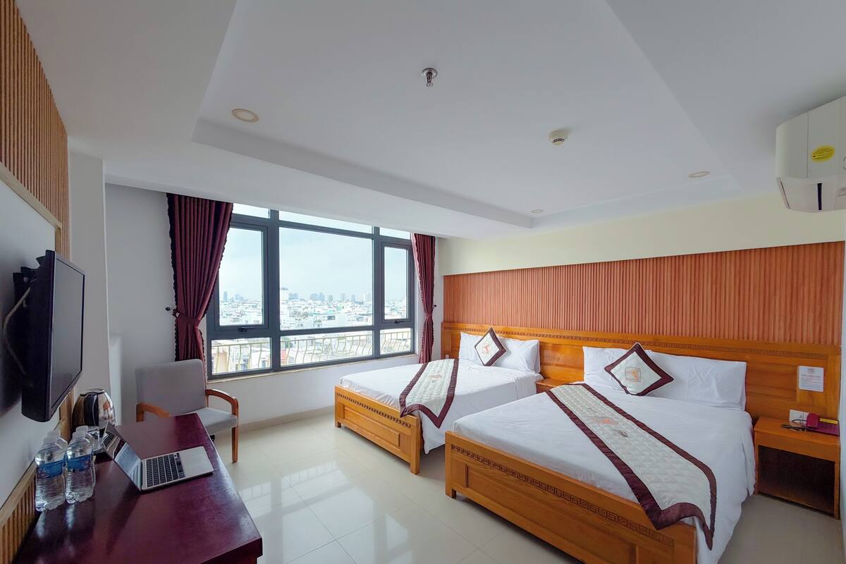 A hotel room with two beds, a desk with a laptop, a television, and a large window with a city view.