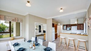 A dining area with a table set for four, a kitchen with bar stools, and a view through a window. - Charming on Cambridge (Napier)