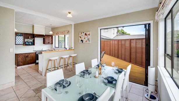 A modern kitchen with a dining area, featuring a large island, white cabinetry, and a view of the backyard. - Charming on Cambridge (Napier)