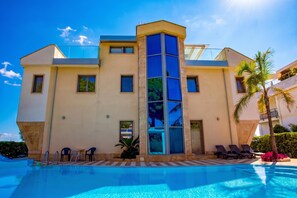 Un hotel modern amb una piscina, múltiples balcons i un cel blau clar.