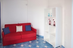 Un sofá rojo con dos almohadones, una estantería blanca con adornos y suelo con azulejos azules.