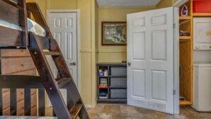 Una habitación con una escalera de madera, una puerta blanca, un estante con cajas de almacenamiento y un cuadro en la pared.