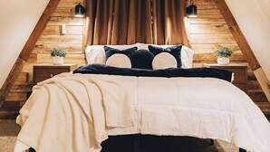 Een slaapkamer met schuine plafonds, een bed met witte beddengoed, twee nachtkastjes met planten, en een raam met gordijnen.