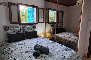 Ein Schlafzimmer mit zwei Betten, jeweils mit blumigem Bettbezug und dunklen Kissen. Es gibt ein Fenster mit blauem Rollo und einen Spiegel über den Betten.