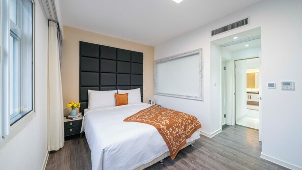 Et moderne hotelværelse med en stor seng, et natbord med lampe, og udsigt til badeværelset gennem en åben dør.