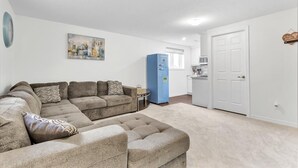 Ein Wohnzimmer mit einem Ecksofa, einem Pouf, einem blauen Kühlschrank und einer weißen Tür.