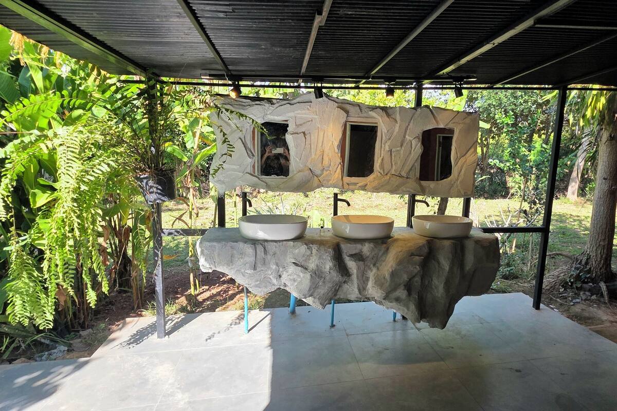 A stone sink with three bowls under a rustic shelter, surrounded by lush greenery.