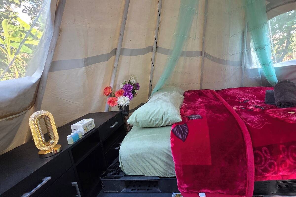 A tent with a bed, a bedside table with a lamp, and a vase with flowers.