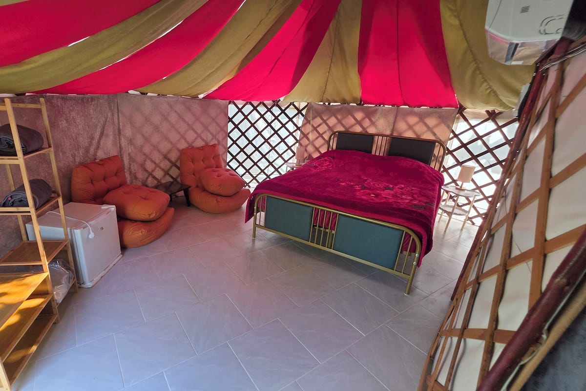 A compact room with a bed, a small desk, and a chair. There is a ladder and a shelf unit.