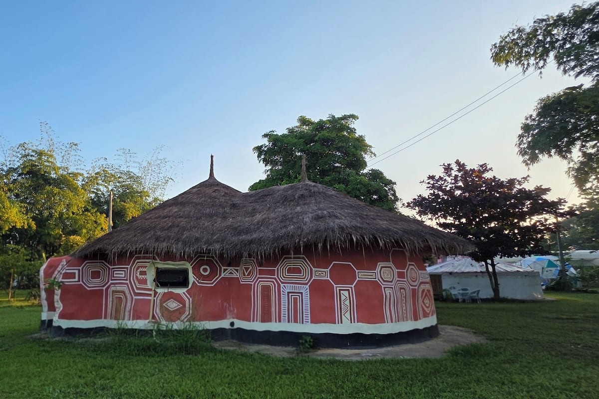 A traditional hut with a thatched roof and red walls, surrounded by greenery.