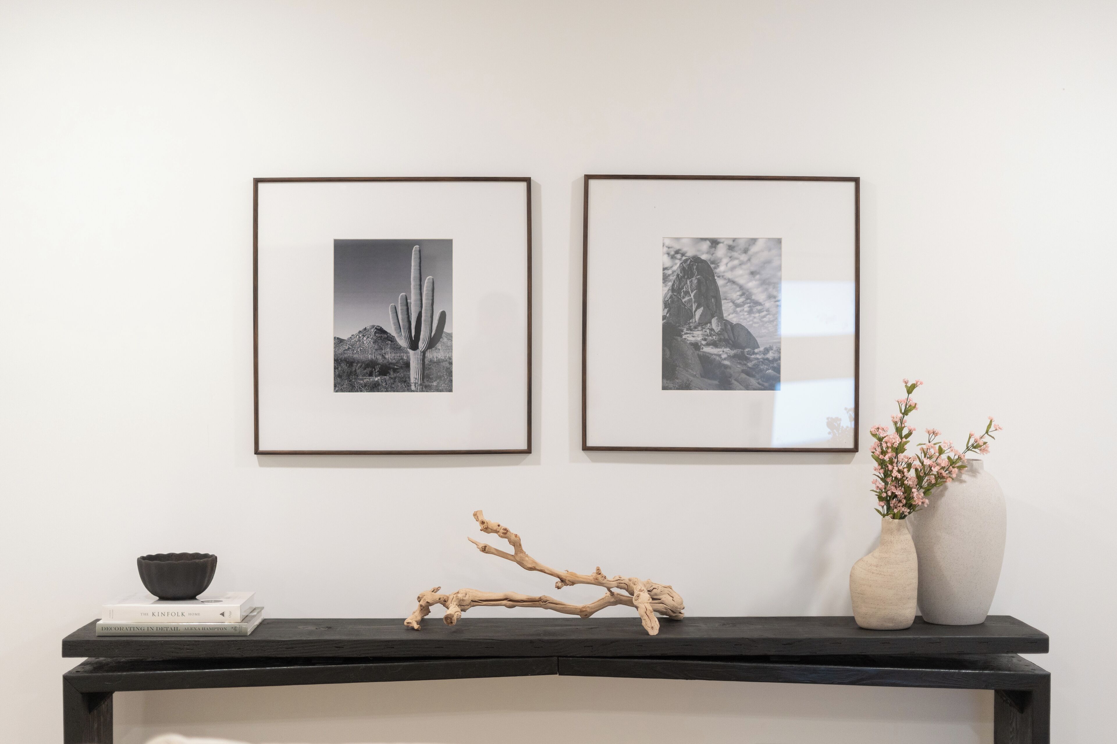 Una pared con dos fotografías blanco y negro enmarcadas, un estante pequeño con un cuenco y libros, y un florero con flores.