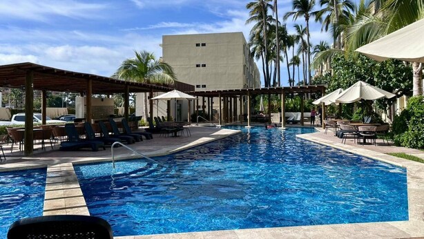 Piscina com espreguiçadeiras e guarda-sóis, rodeada por palmeiras e um edifício ao fundo.