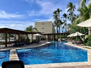A swimming pool with lounge chairs and umbrellas, surrounded by palm trees and a building in the background. - Departamento en Oceanna Condos del Mar Cerritos (Mazatlán)