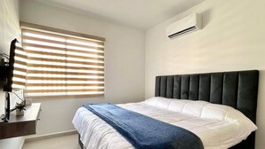 Quarto com cama, mesa de cabeceira, janela com persianas e aparelho de ar condicionado.