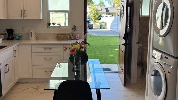 Een moderne keuken met witte kasten, een glazen tafel en een zwarte stoel. Er is een raam met uitzicht op een groen gazon en een geparkeerde auto.