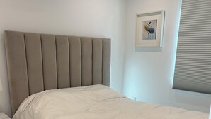 Een slaapkamer met een bed, een nachtkastje, een lijst met een afbeelding aan de muur en een raam met jaloezieën.