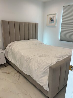 A bedroom with a bed, a nightstand, a framed picture on the wall, and a window with blinds. - Cozy 1-bedroom apartment in Los Angeles with AC (Los Angeles)