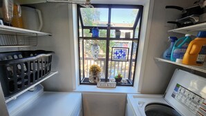 A compact laundry room with a window, a washing machine, a dryer, and shelves with cleaning supplies. - In the trendy, funky & walkable South Congress area — 2-bedroom condo in Austin! (Austin)
