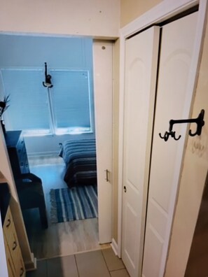 Een smalle gang die leidt naar een slaapkamer met een bed, een klein tafeltje en een raam.