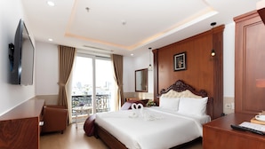 A hotel room with a large bed, a wooden headboard, a flat-screen TV mounted on the wall, a desk with a keyboard, and a view of the city through the window. - Quan Cuong Hotel (Da Nang)