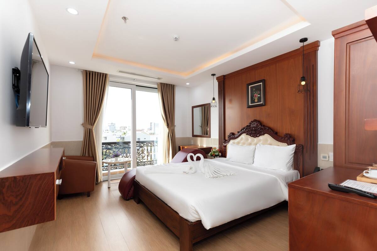 Een hotelkamer met een groot bed, een houten hoofdbord, een flatscreen televisie aan de muur, een bureau met toetsenbord en uitzicht op de stad.