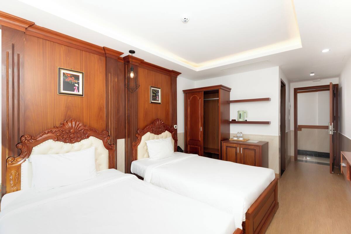 Hotelkamer met twee bedden, houten hoofdborden, een garderobe, een klein tafeltje met lamp en een douche.