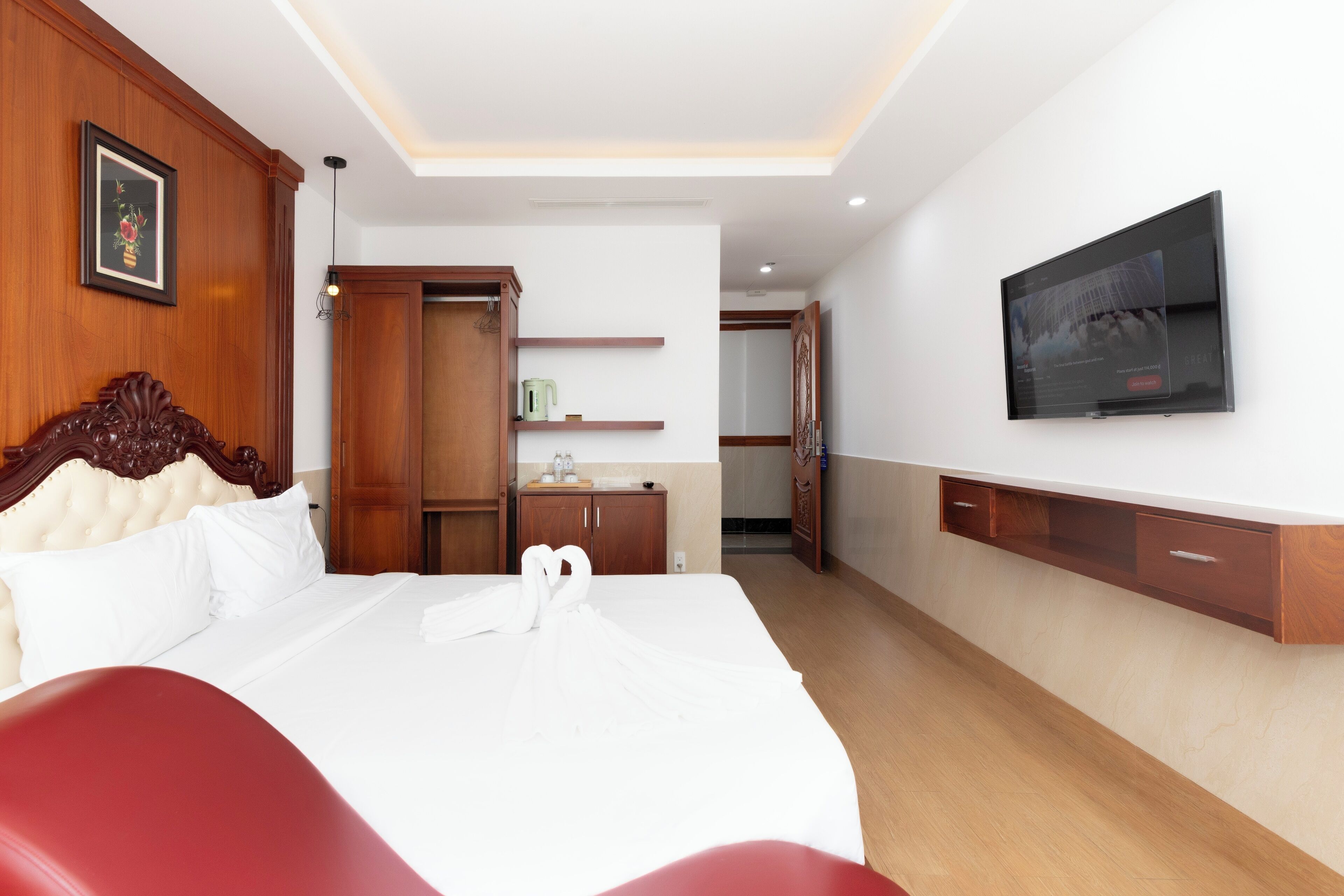 A hotel room with a large bed, wooden wardrobe, a television mounted on the wall, and a small kitchenette area.