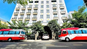 A multi-story building with a central entrance, surrounded by trees and two buses parked in front. - Quan Cuong Hotel (Da Nang)