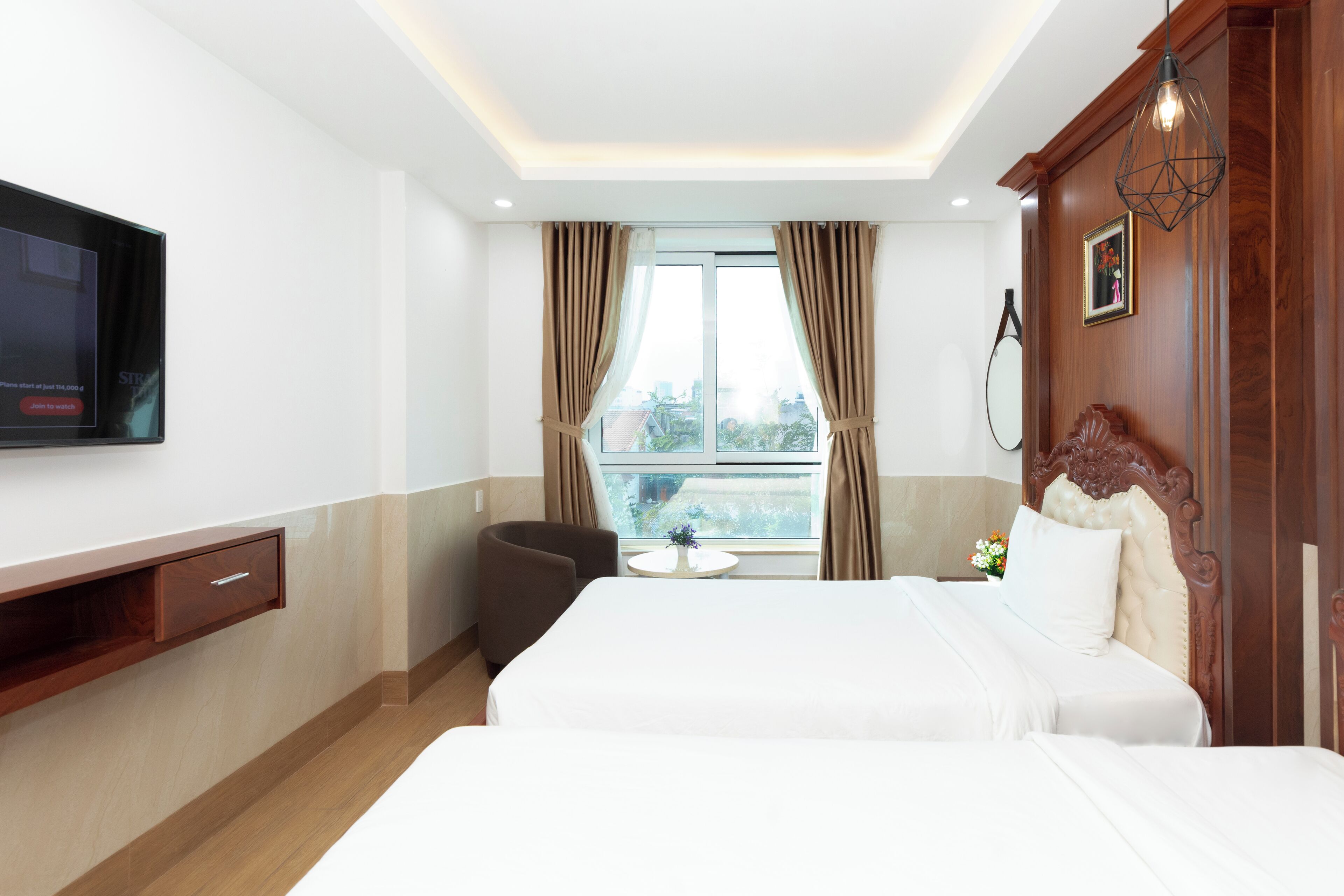 A hotel room with a bed, a TV mounted on the wall, a small table with a vase, and a window with curtains.