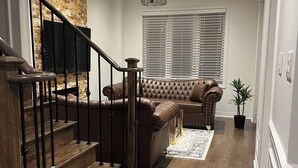 A modern living room with a wooden staircase, a brown leather sofa, a chandelier, and a large window with blinds. - Warm & Stylish 4BR Home | 3BA • Near Toronto (Ajax)