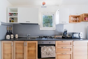A modern kitchen with a gas stove, oven, and microwave. There are wooden cabinets, a window with a view of trees, and a shelf with spices and jars. - Pajara Fazzadio by Vivimy (Presicce)