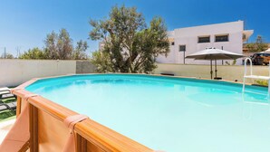 Ein Schwimmbecken mit einer Holzterrasse, einem weißen Sonnenschirm und einem Gebäude im Hintergrund.