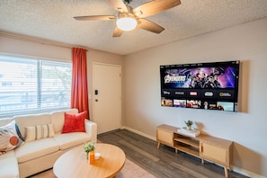 A living room with a ceiling fan, a flat-screen TV displaying a movie, a sofa, a coffee table, and a small wooden side table.