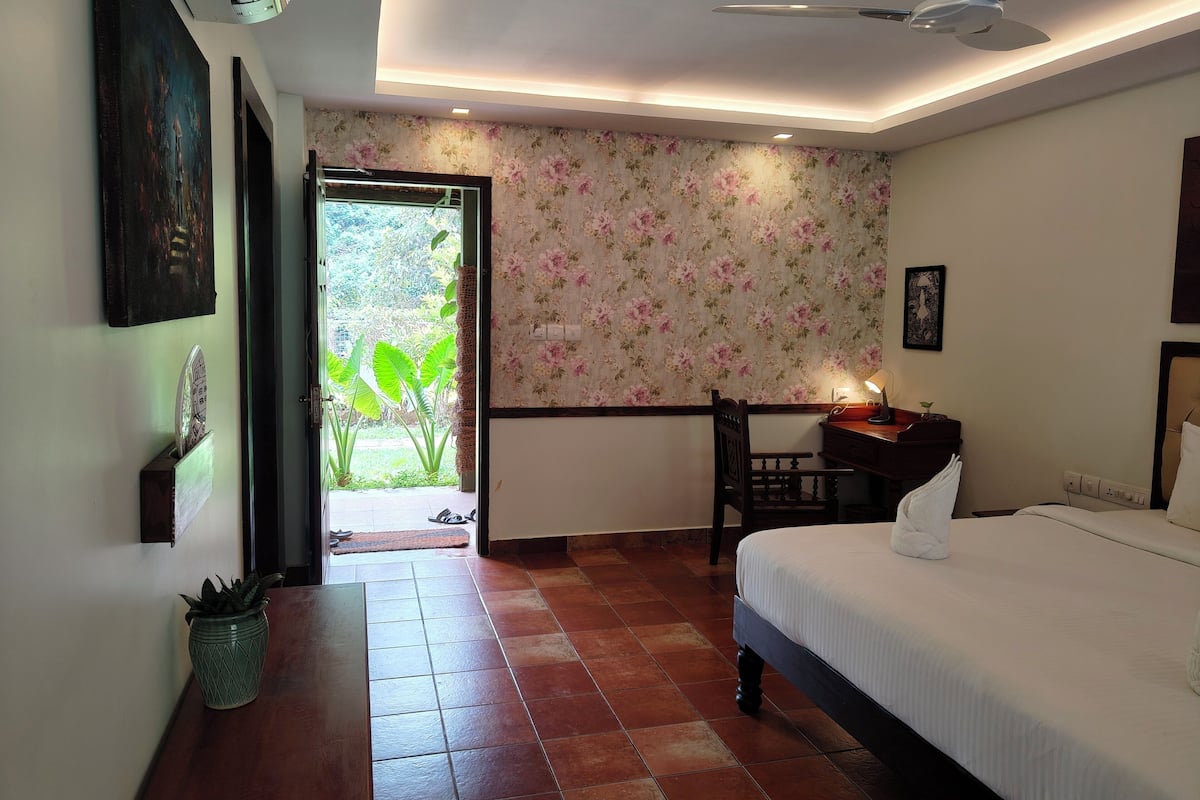 A hotel room with a bed, a desk, a chair, a potted plant, and a view of the outdoors.