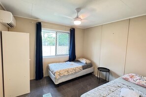 Un dormitorio con cama, una mesita pequeña, una ventana con cortinas, un equipo de aire acondicionado y un ventilador de techo.
