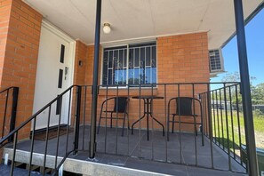 Un espacio exterior cubierto con pared de ladrillo, una ventana con rejas de metal, dos sillas metálicas y una mesa pequeña.