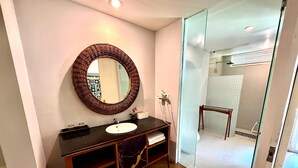 A modern bathroom with a round mirror, dark wooden vanity, and a glass shower enclosure. - Dealton Hotel Sanur (Denpasar)