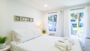 A bedroom with a bed, a bedside table with a lamp, a potted plant, and a window with curtains. - One Block Walk to Beach - Get Your Toes in the Sand in Minutes!  Covered Deck, Garden Views & Fun (Folly Beach)