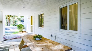 A wooden picnic table with benches and a potted plant in the center, set on a porch with white siding and large windows. - One Block Walk to Beach - Get Your Toes in the Sand in Minutes!  Covered Deck, Garden Views & Fun (Folly Beach)