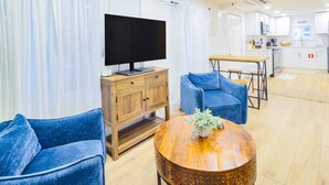 A modern living room with a TV, blue sofas, a wooden coffee table, and a kitchen in the background. - One Block Walk to Beach - Get Your Toes in the Sand in Minutes!  Covered Deck, Garden Views & Fun (Folly Beach)