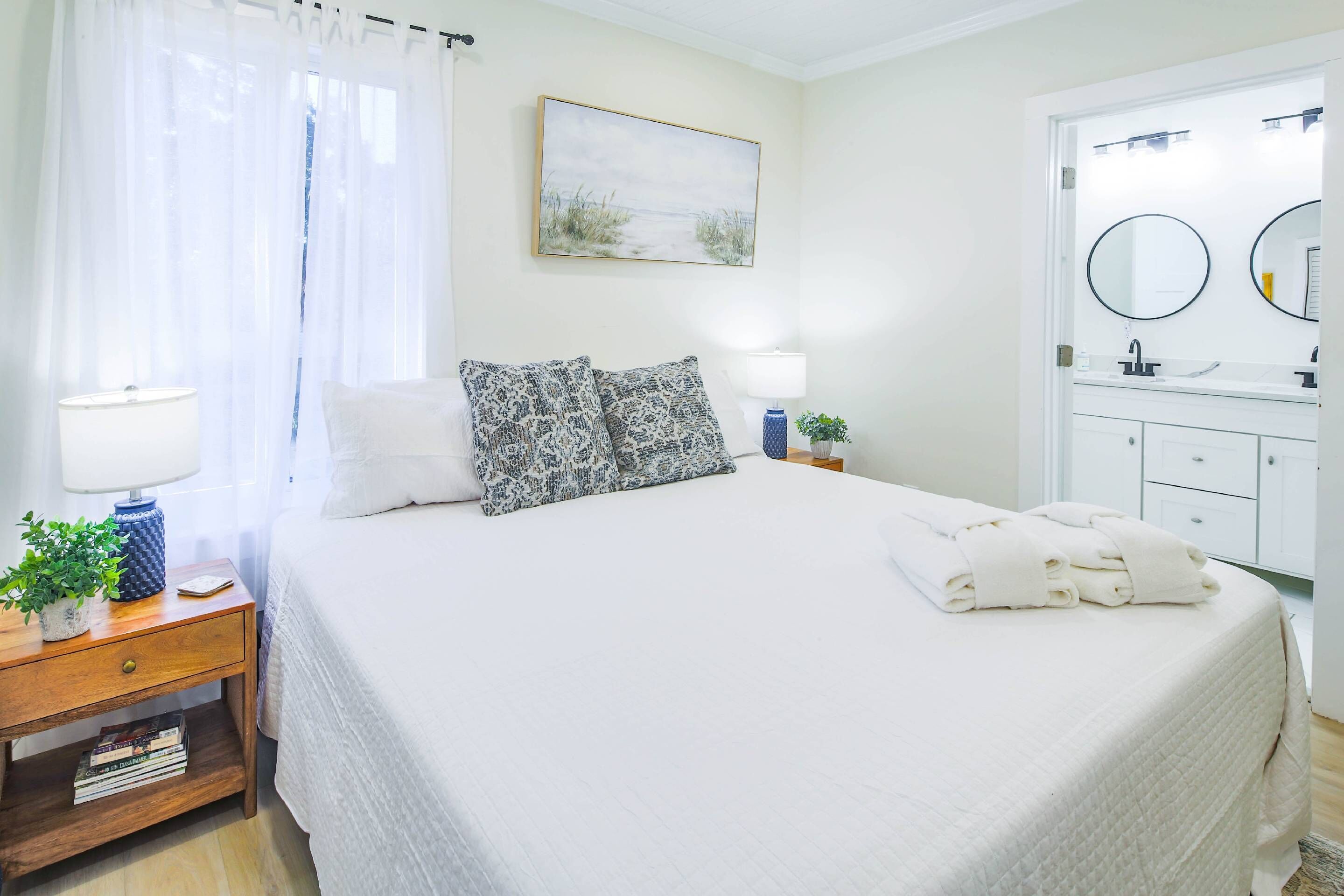 A neatly made bed with white bedding, a bedside table with a lamp, and a wall-mounted painting.