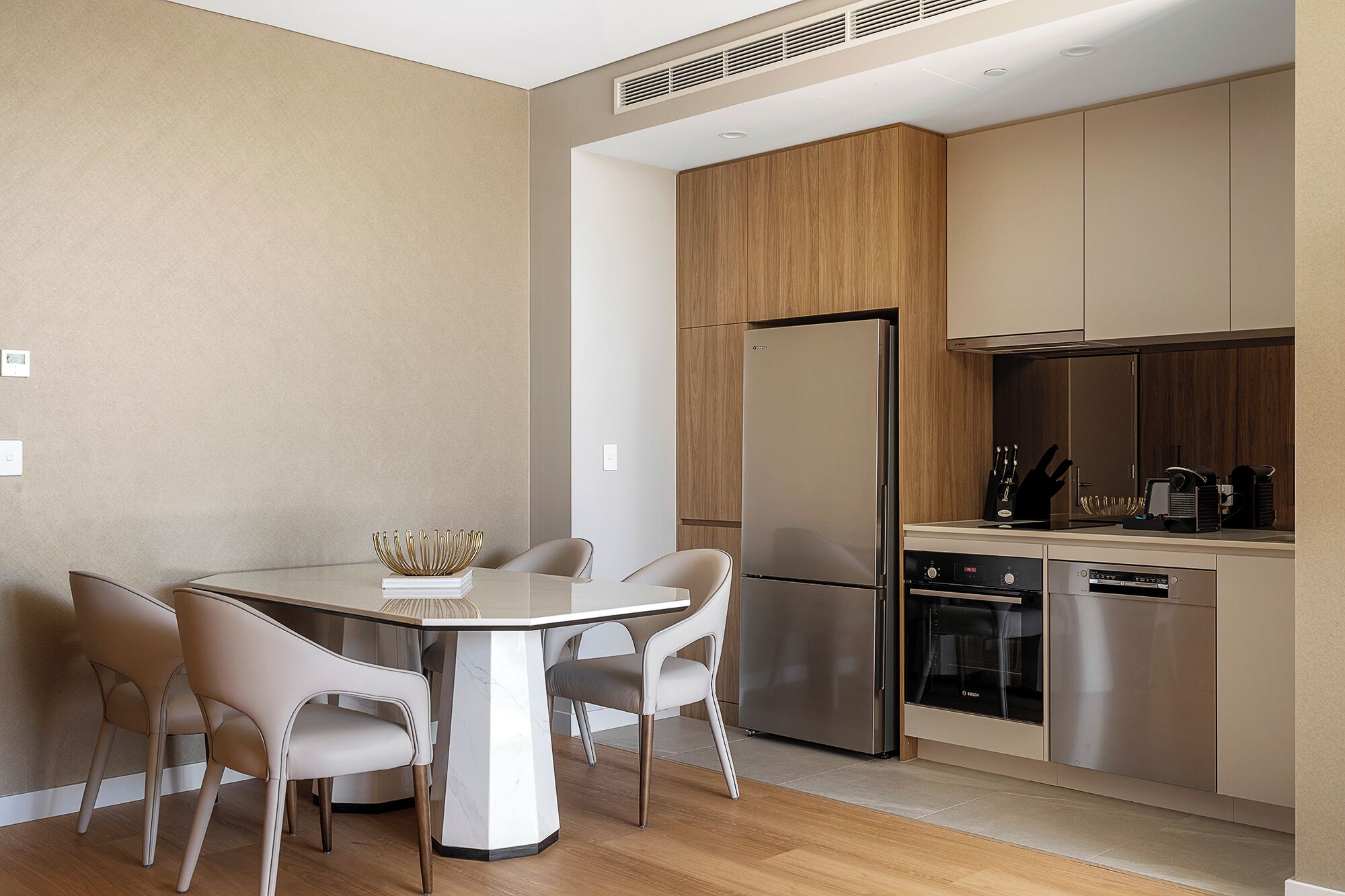 A modern kitchen with a stainless steel refrigerator, oven, and dishwasher. There is a dining table with four chairs and a decorative bowl on it.