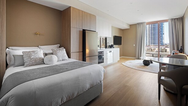 Ein modernes Hotelzimmer mit einem großen Bett, einer Küchenzeile, einem Essbereich und einem Balkon mit Blick auf die Stadt.