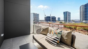 Ein Balkon mit einer Couch, Kissen und Blick auf eine Stadtlandschaft mit Gebäuden und Kränen.