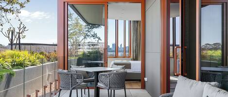 A balcony with a dining table and chairs, a sofa, and a view of a cityscape through large windows.