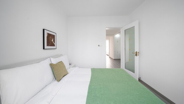 Habitación de hotel con cama, manta verde, ropa de cama blanca, un cuadro en la pared y una puerta que da a otra habitación.
