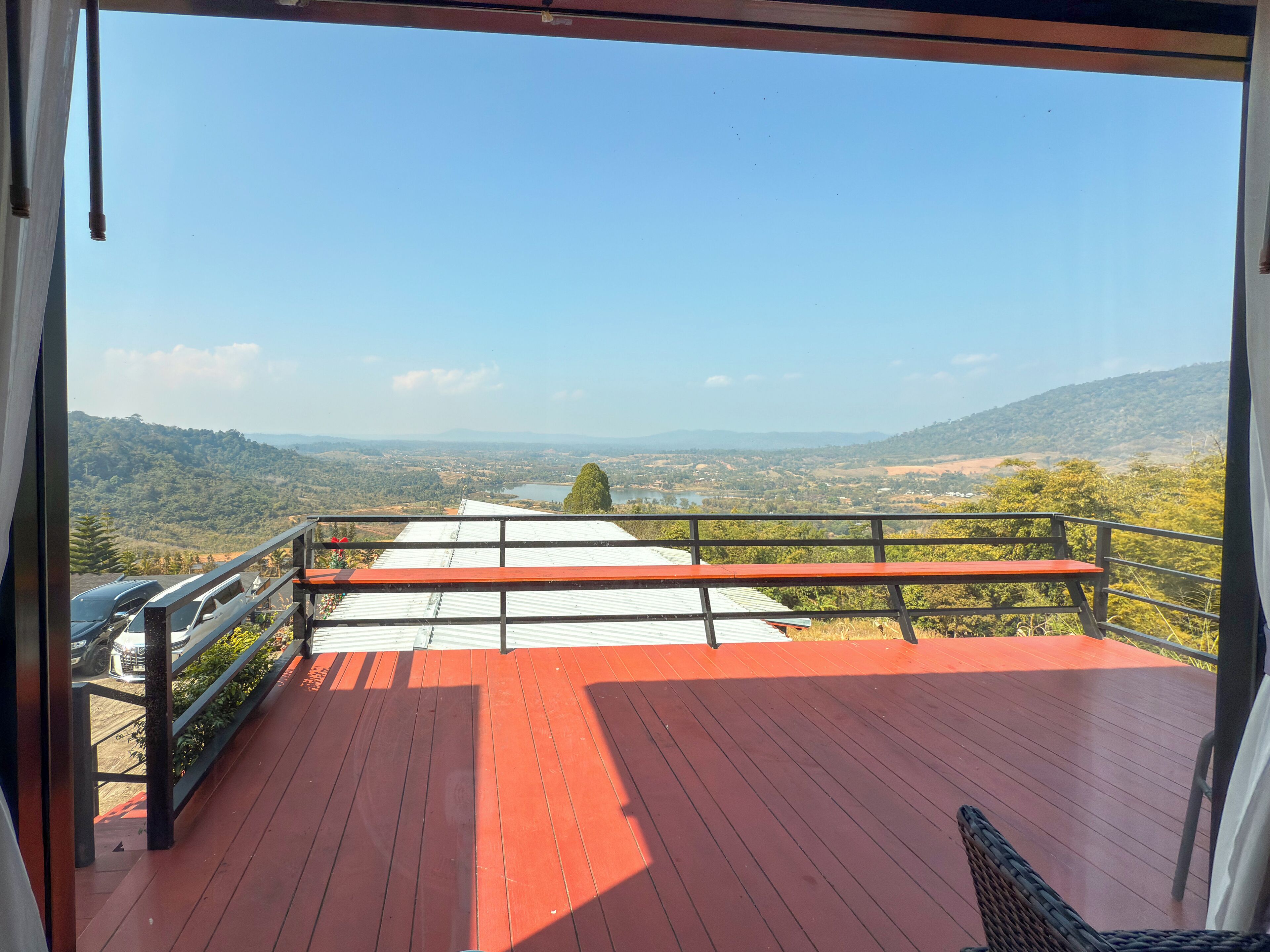 A balcony with a red floor and metal railing, overlooking a scenic landscape with hills and a river.
