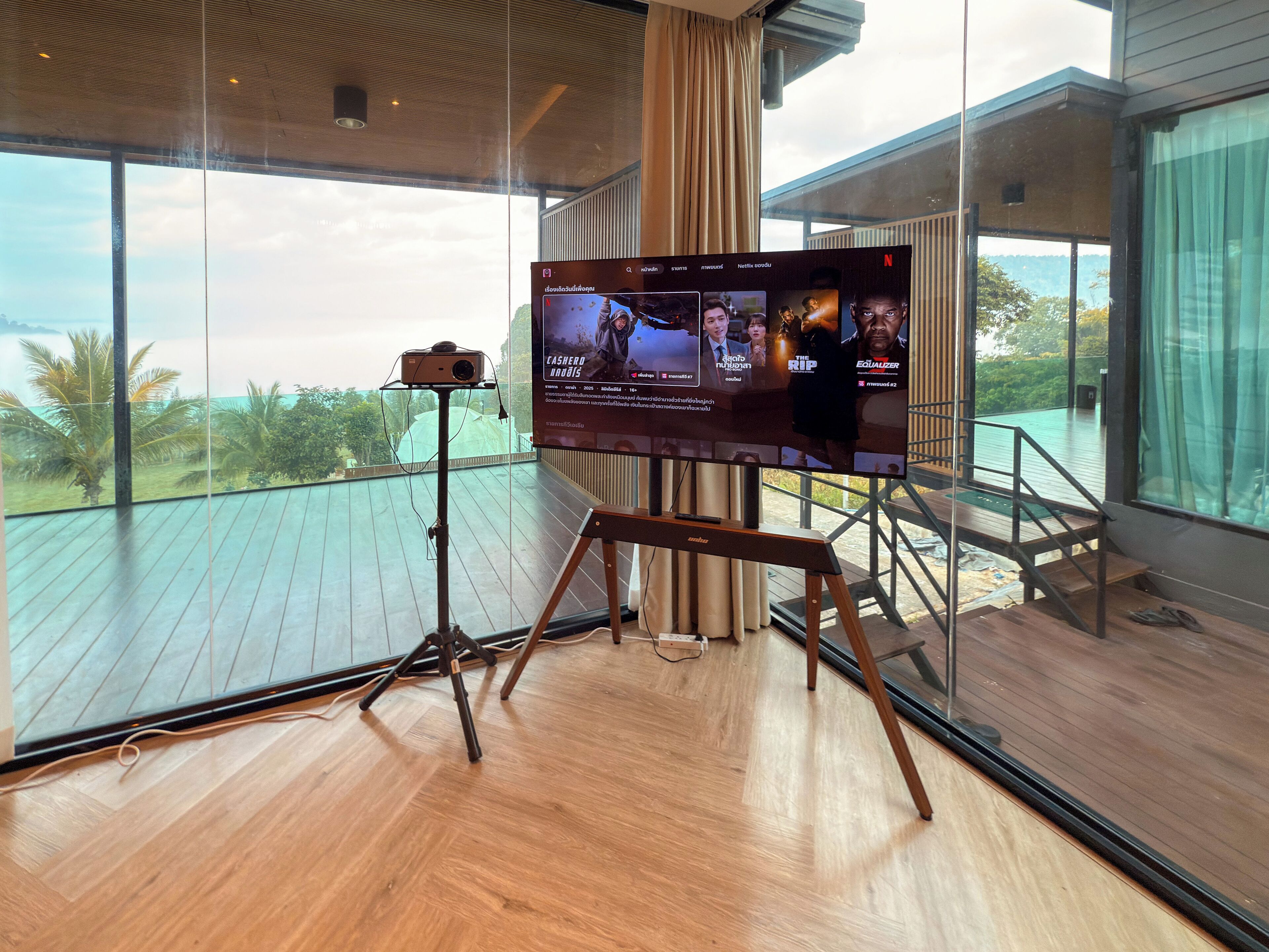 Quarto moderno com uma tela grande exibindo um filme, piso de madeira e varanda com vista para o mar.