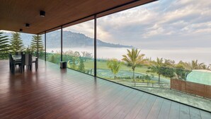 Un balcón amplio con piso de madera y una mesa con sillas, con vistas a un paisaje exuberante con árboles y una cadena montañosa distante.