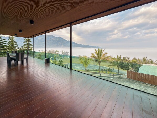 A spacious balcony with a wooden floor and a table with chairs, overlooking a lush landscape with trees and a distant mountain range. - Laguna Khaokho (Khao Kho)